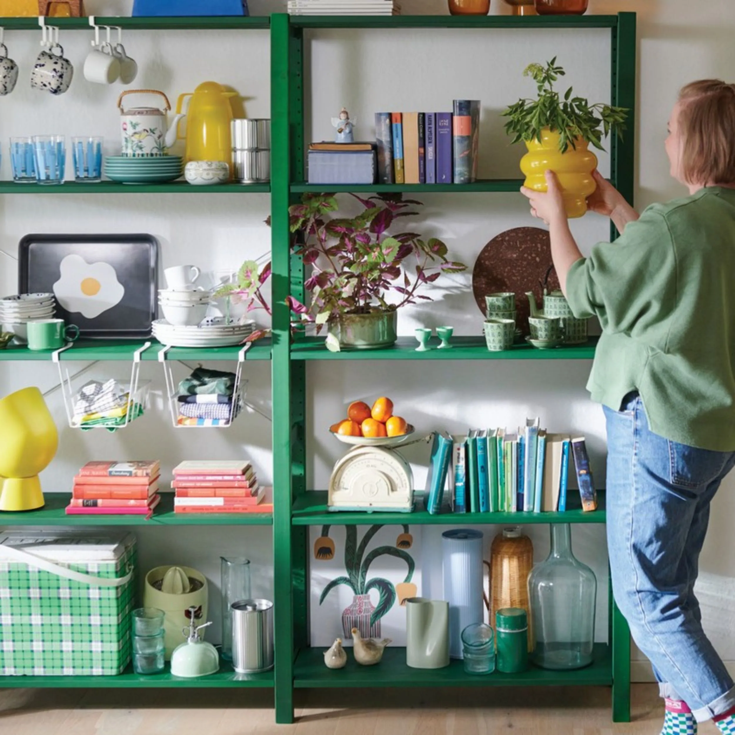 Green IKEA shelf with dishes and books, a woman placing a yellow pot